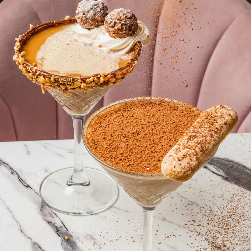 Two dessert cocktails on a marble table, one with nuts and chocolates, the other with cocoa and a ladyfinger.