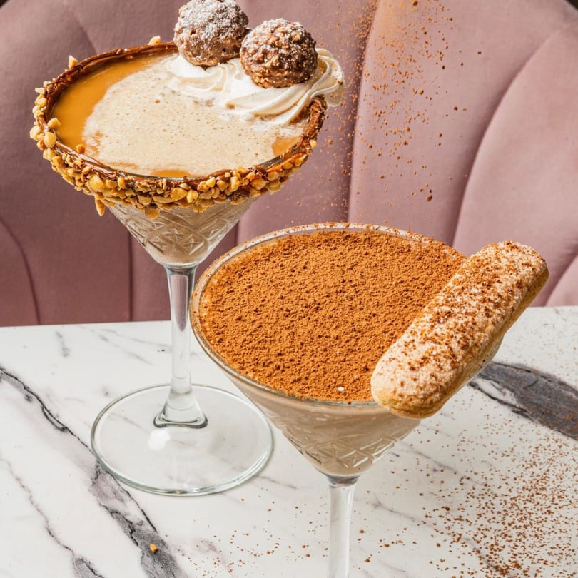 Two dessert cocktails on a marble table, one with nuts and chocolates, the other with cocoa and a ladyfinger.
