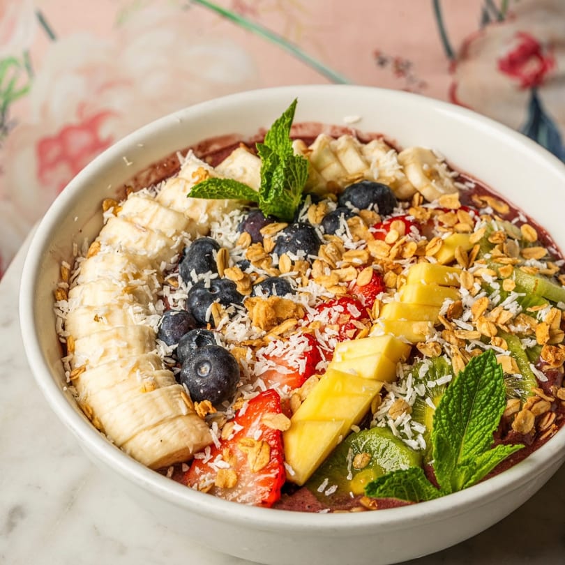 A vibrant acai bowl topped with bananas, blueberries, strawberries, mango, kiwi, granola, and mint.