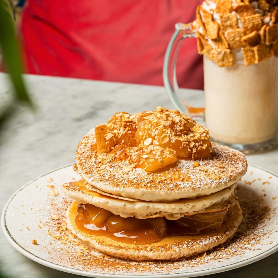 Stack of pancakes with apple compote, powdered sugar, and nuts, next to a milkshake.