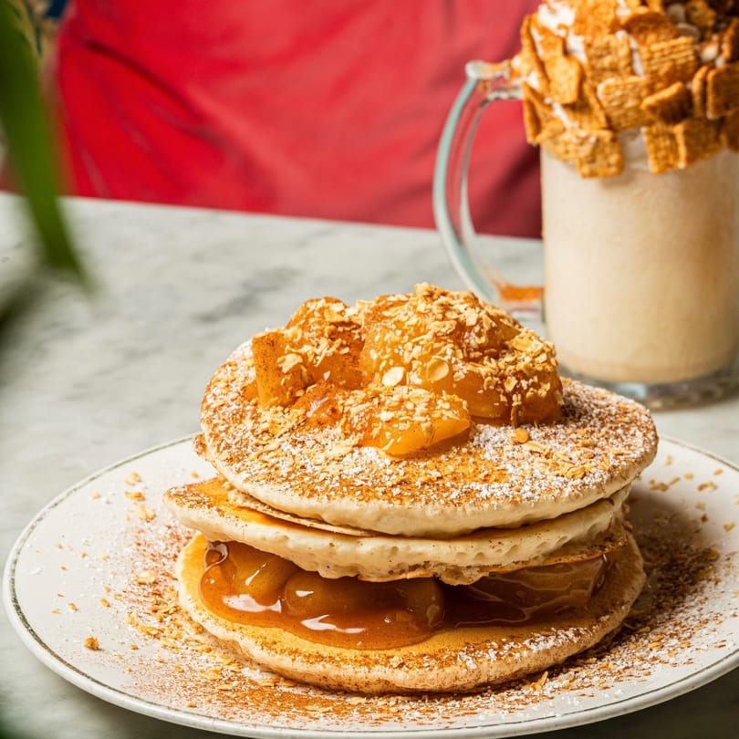 Stack of pancakes with apple compote, powdered sugar, and nuts, next to a milkshake.