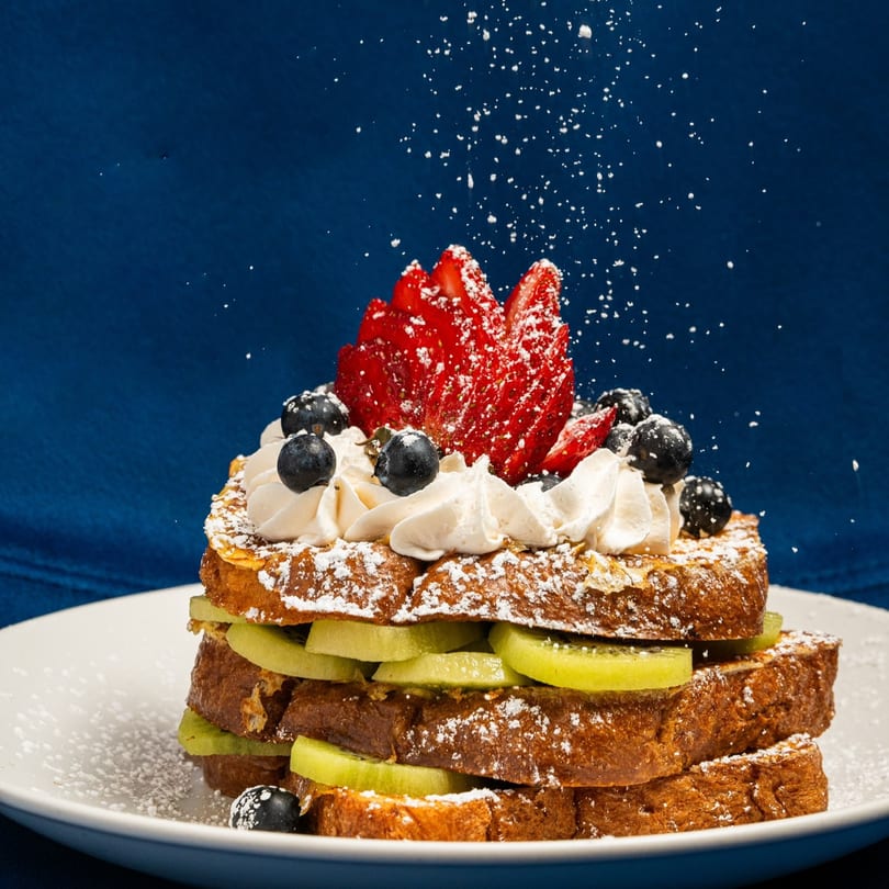 Stacked French toast with kiwi, whipped cream, blueberries, strawberries, and powdered sugar falling.